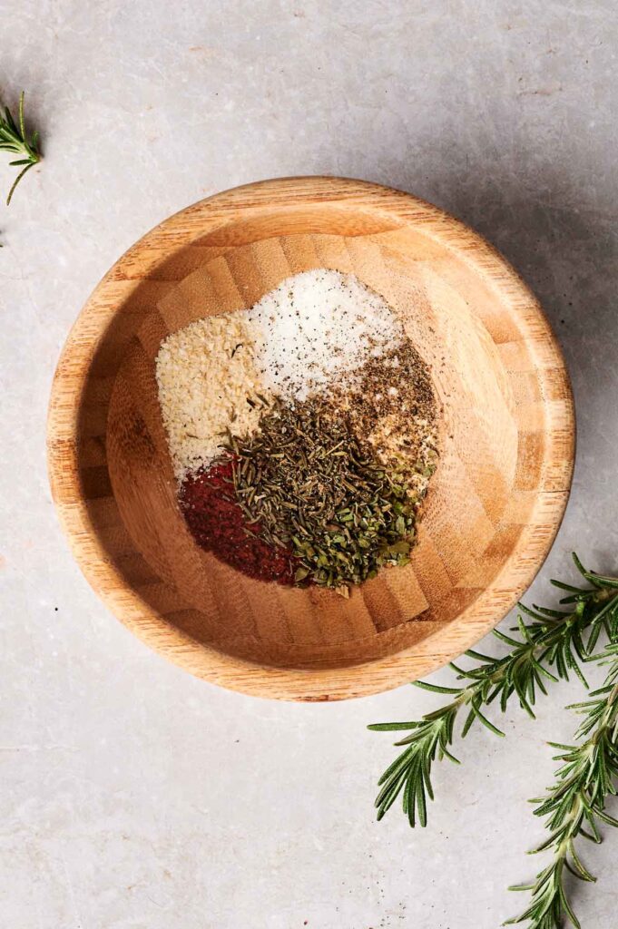 A wooden bowl containing a mixture of spices and herbs, with a sprig of rosemary on the side.