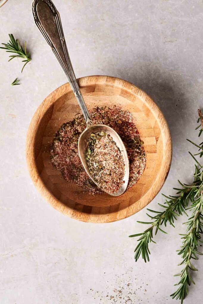 Wooden bowl with mixed spices and a spoon inside. Sprigs of rosemary are placed beside the bowl on a light textured surface.