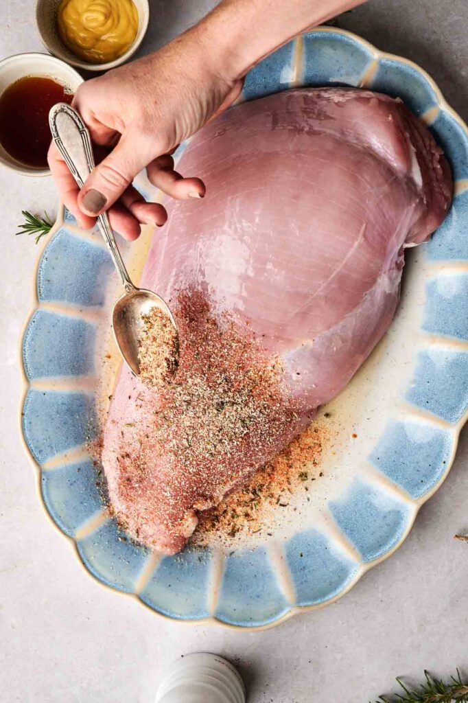 A hand sprinkles seasoning onto a raw turkey breast on a blue-rimmed platter, with mustard and sauce bowls nearby.