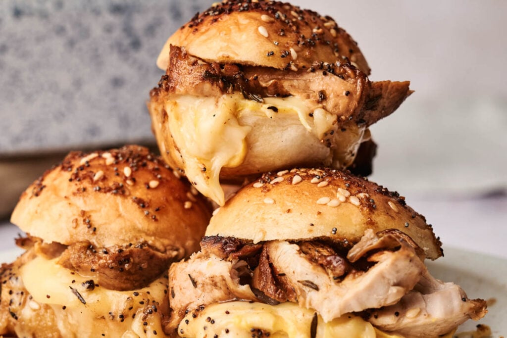 Three chicken, turkey, and cheese sliders with seasoned, sesame-topped buns are stacked on a plate. Melted cheese and roasted chicken and turkey are visible between the buns.