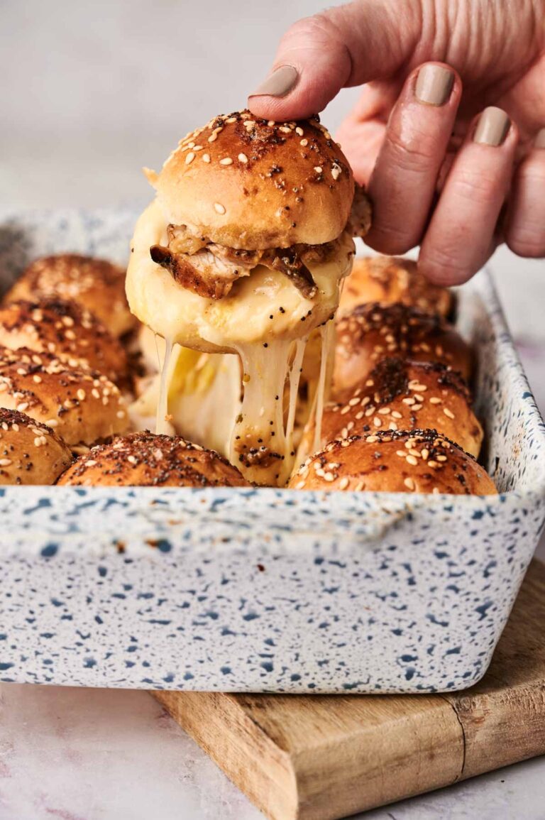 A hand lifts a cheesy turkey slider from a speckled baking dish, filled with similar sliders topped with sesame seeds.