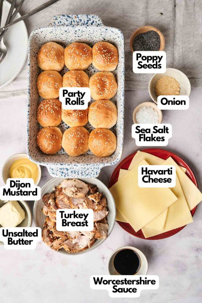 Top view of ingredients for turkey sliders: party rolls, poppy seeds, onion, sea salt flakes, unsalted butter, turkey breast, Worcestershire sauce, Havarti cheese, and Dijon mustard.