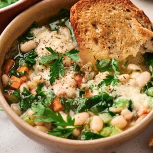 A bowl of white bean soup with carrots, greens, and cheese, topped with a slice of toasted bread.