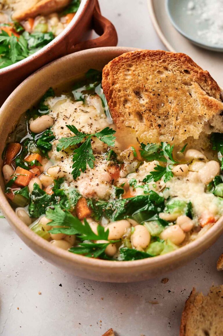 A bowl of white bean soup with carrots, greens, and cheese, topped with a slice of toasted bread.