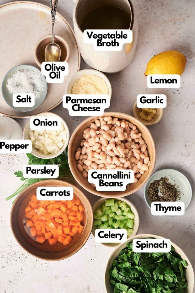 Ingredients for a white bean soup are laid out on a table: vegetable broth, olive oil, lemon, salt, pepper, garlic, onion, parmesan cheese, parsley, cannellini beans, carrots, celery, thyme, and spinach.