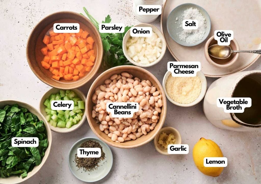 Bowls and plates holding ingredients for white bean soup: chopped carrots, celery, spinach, onion, cannellini beans, parsley, thyme, garlic, salt, pepper, parmesan cheese, olive oil, vegetable broth, and a lemon.