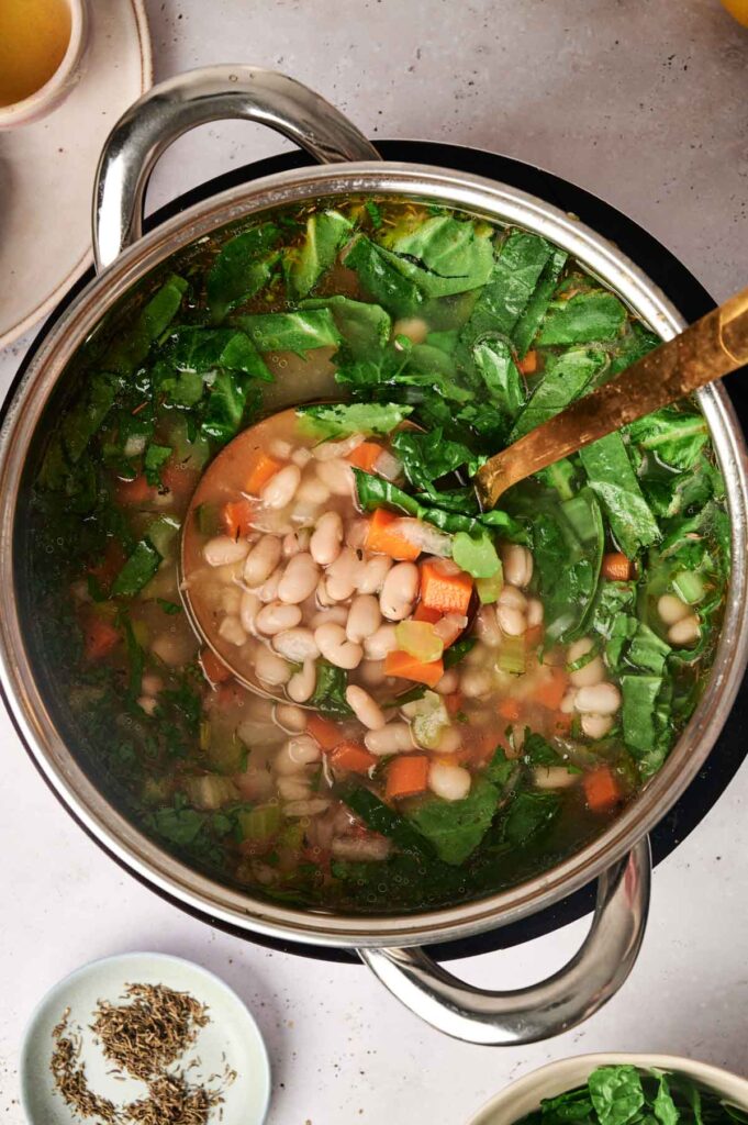A pot of hearty white bean soup brimming with leafy greens, carrots, and savory broth. A ladle is partially submerged, ready to serve comfort in a bowl.