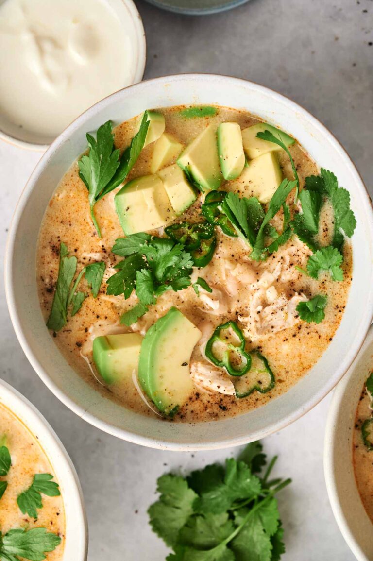 Bowl of creamy white chicken chili topped with avocado slices, cilantro, parsley, and green chili, served with a side of sour cream—perfect for those who love the flavors of an air fryer twist.