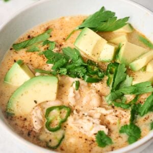 A bowl of white chicken chili soup, brimming with creamy goodness, is topped with tender chicken pieces, fresh avocado slices, and a sprinkle of green herbs.