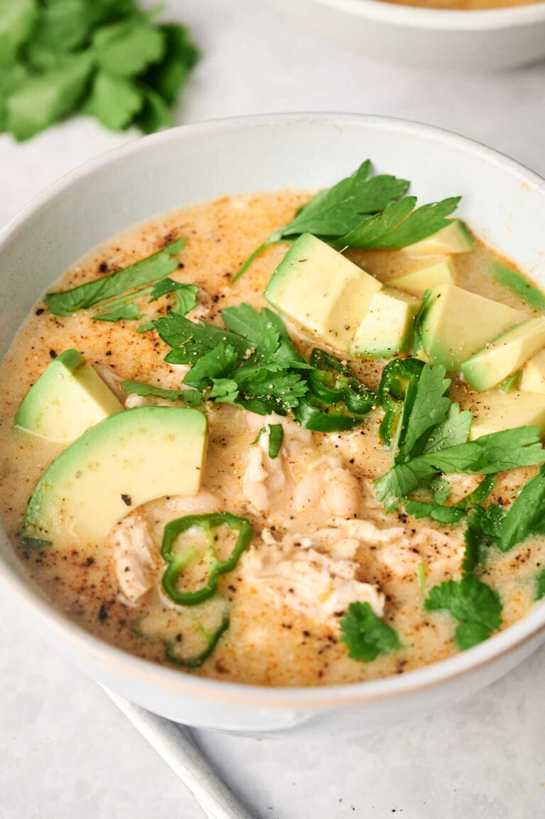 A bowl of white chicken chili soup, brimming with creamy goodness, is topped with tender chicken pieces, fresh avocado slices, and a sprinkle of green herbs.