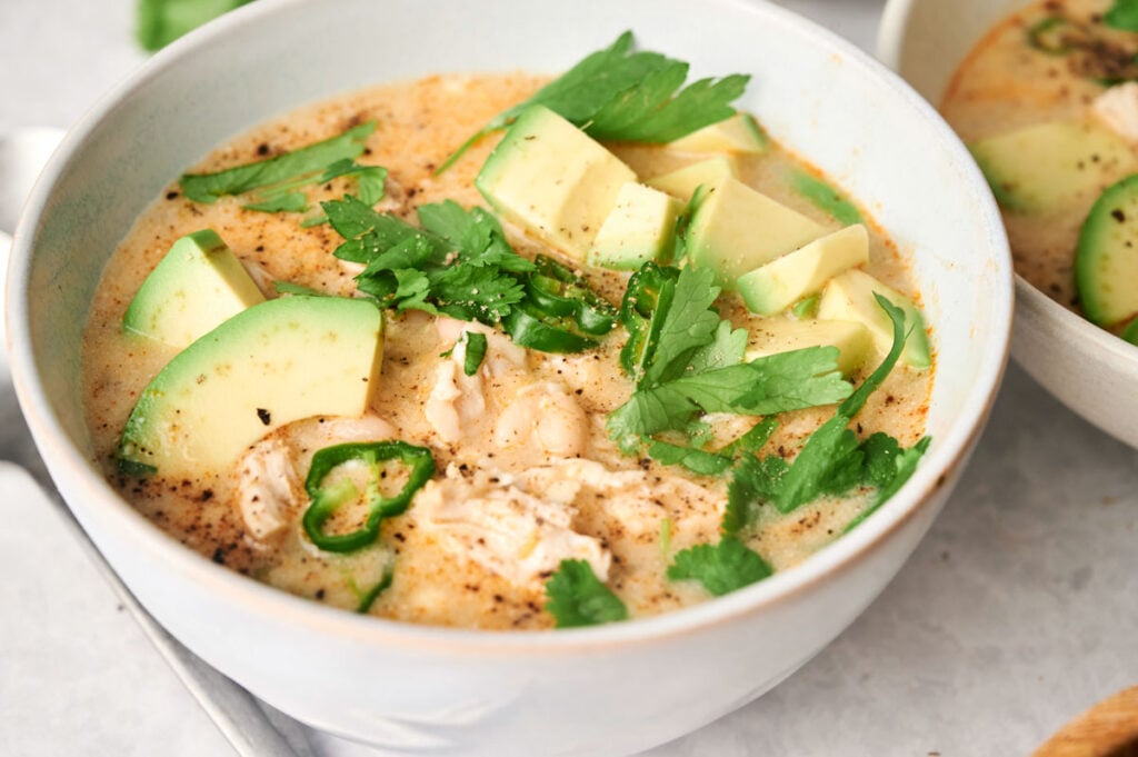 A bowl of creamy White Chicken Chili topped with sliced avocado, fresh cilantro, jalapeño, and tender pieces of chicken.