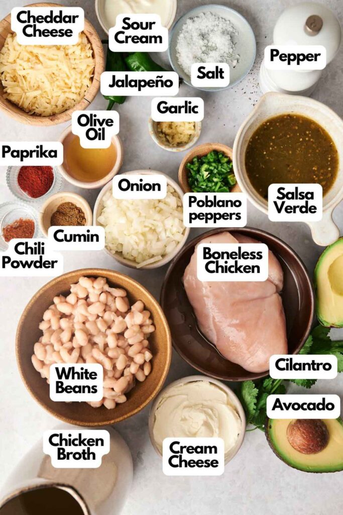 An assortment of ingredients for a white chicken chili recipe, including raw chicken, spices, vegetables, beans, cheese, and avocado, arranged on a countertop.