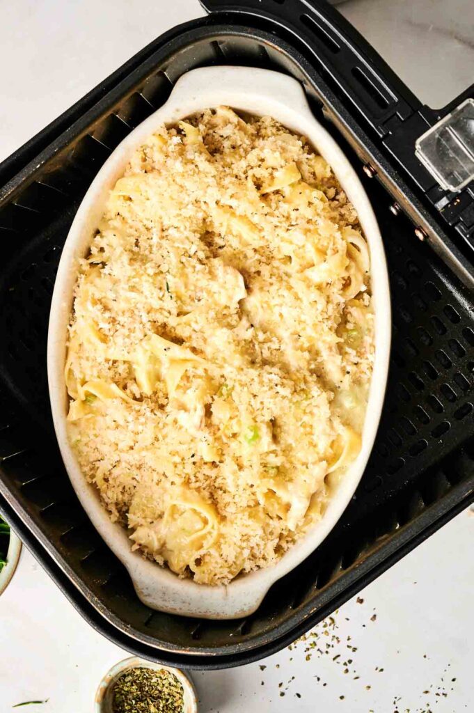 Oval dish with creamy Air Fryer Chicken Noodle Casserole topped with breadcrumbs inside an air fryer basket. Herbs visible on the side.
