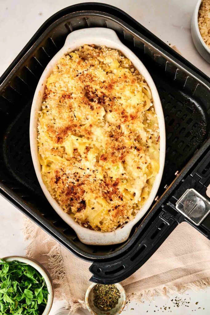 A delicious Air Fryer Chicken Noodle Casserole features a golden breadcrumb topping in a white dish, sitting perfectly inside an air fryer basket. Nearby, small bowls hold chopped herbs and seasoning to enhance the flavorful experience.