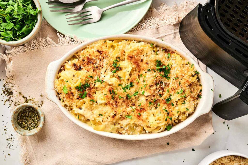A casserole dish features baked pasta topped with breadcrumbs and herbs, reminiscent of an Air Fryer Chicken Noodle Casserole. Nearby, a green plate with forks sits beside a bowl of chopped herbs and a spice container.