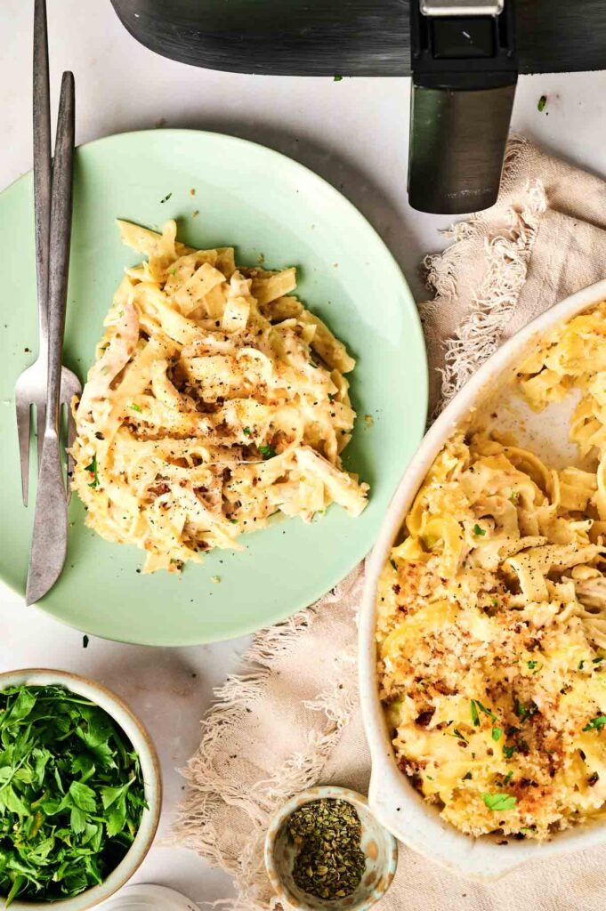 A plate of creamy pasta with herbs and black pepper sits next to a serving dish, reminiscent of an Air Fryer Chicken Noodle Casserole, alongside bowls of parsley and seasoning on a cloth napkin.