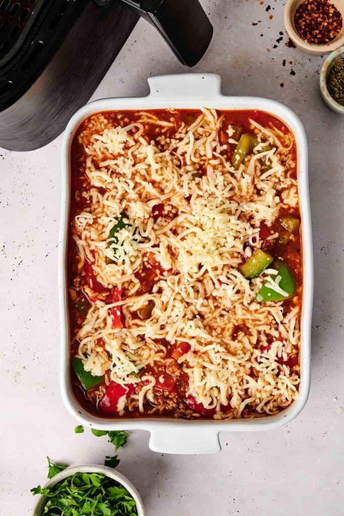 A white baking dish filled with an unbaked air fryer stuffed pepper casserole featuring tomato sauce, sliced vegetables, ground meat, and shredded cheese. Nearby are small bowls of spices and chopped herbs.