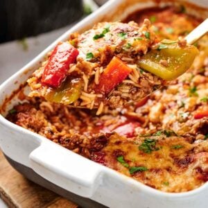 A serving of Air Fryer Stuffed Pepper Casserole with rice, bell peppers, and melted cheese is being lifted from a white baking dish.