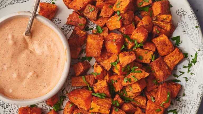 A plate of roasted sweet potato cubes garnished with herbs, accompanied by a bowl of creamy dipping sauce with a spoon.