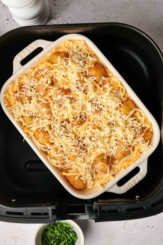 An air fryer tater tot breakfast casserole sits invitingly in the basket, with crispy tots smothered in melted cheese. Nearby, a small bowl of chopped herbs awaits to add a fresh touch to this delicious morning meal.