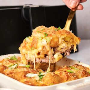 A hand lifts a portion of cheesy, baked air fryer tater tot breakfast casserole with a spatula from a white dish. The delicious casserole is topped with fresh herbs.
