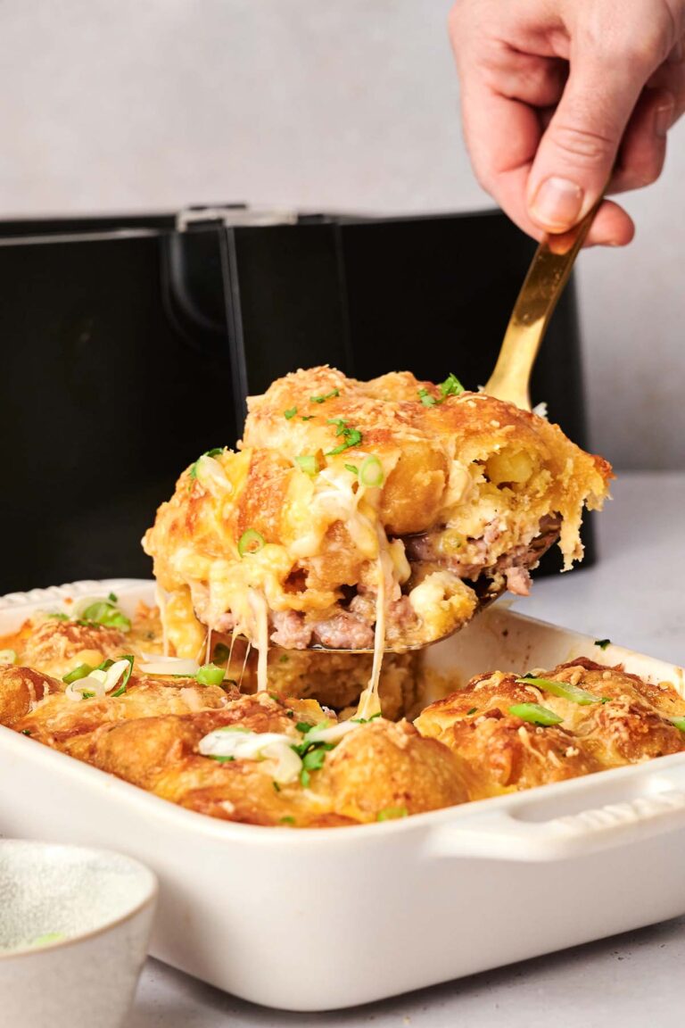 A hand lifts a portion of cheesy, baked air fryer tater tot breakfast casserole with a spatula from a white dish. The delicious casserole is topped with fresh herbs.