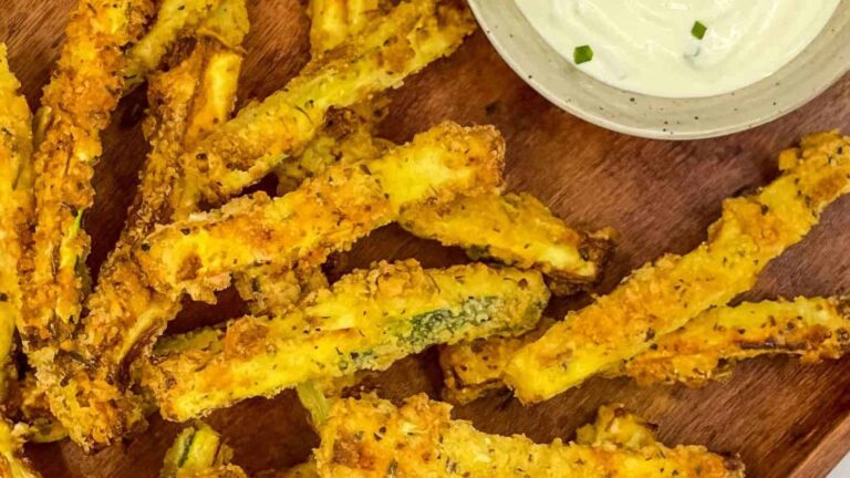 Crispy zucchini fries on a wooden surface next to a small bowl of creamy dipping sauce.