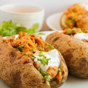 Baked potatoes topped with shredded cheese, sour cream, and herbs sit invitingly on a white plate, with a delicious hint of BBQ chicken stuffed within and sauce in the background.