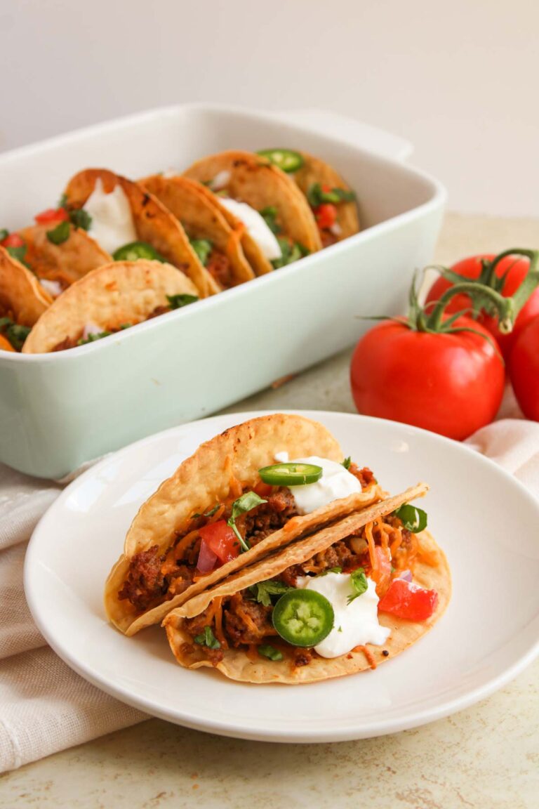 Crispy tacos filled with meat, topped with sliced jalapeños, tomatoes, and sour cream on a white plate. A tray with more tacos and three tomatoes are in the background.