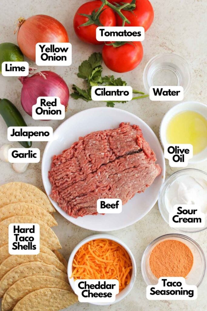 Ingredients for tacos laid out on a table: ground beef, taco shells, cheddar cheese, taco seasoning, sour cream, olive oil, water, tomatoes, cilantro, onions, lime, jalapeno, and garlic.