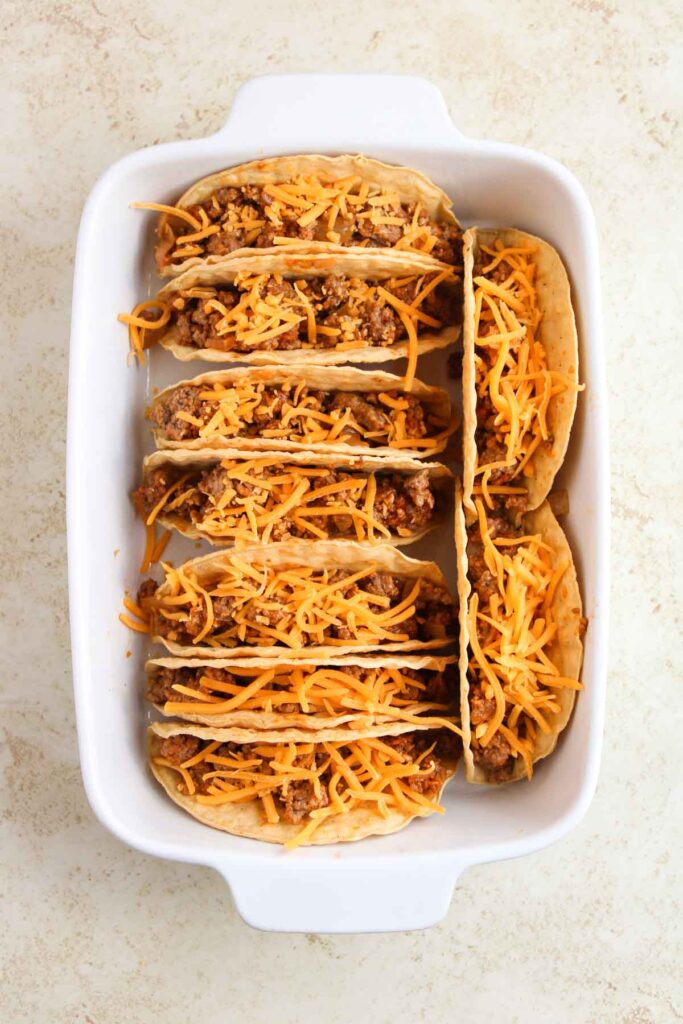 A white baking dish with six tacos filled with ground meat and topped with shredded cheddar cheese.