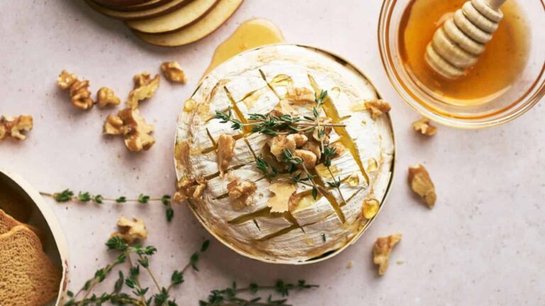 Baked cheese topped with thyme and walnuts, surrounded by honey, thyme sprigs, and crisp bread slices on a light surface.