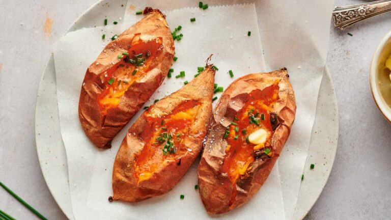 Three baked sweet potatoes topped with chopped chives and garlic on a parchment-lined plate.