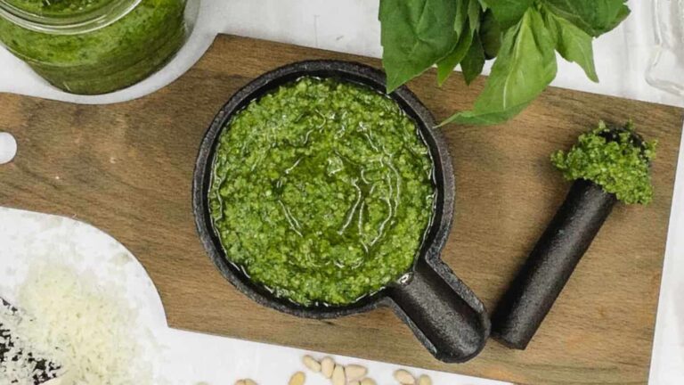 Overhead view of a mortar with green pesto, surrounded by basil leaves, a jar of pesto, grated cheese, pine nuts, and a pestle on a wooden board.