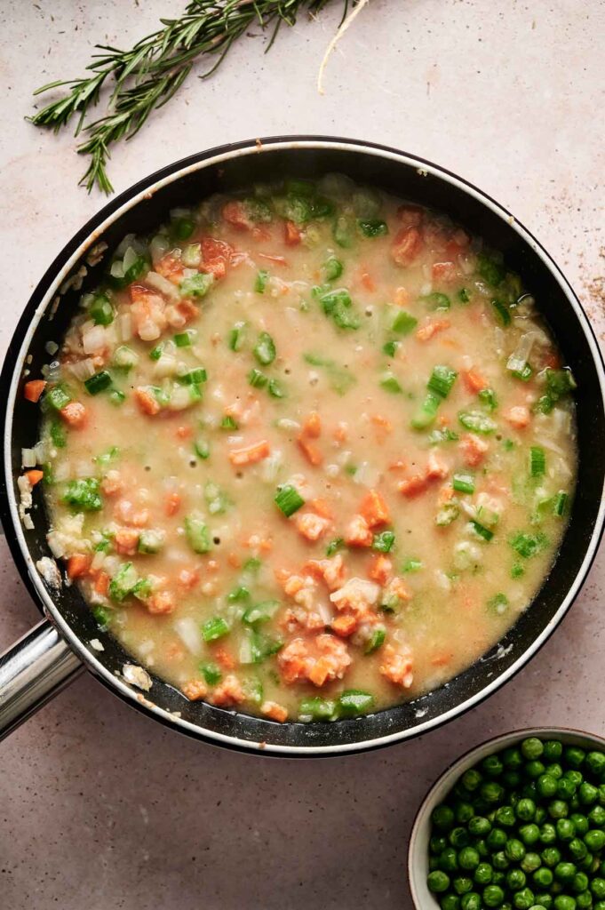 A skillet contains a mixture of diced vegetables and a thick creamy sauce, reminiscent of beef pot pie filling. A small bowl of green peas sits beside it, with sprigs of rosemary adding an aromatic touch in the background.