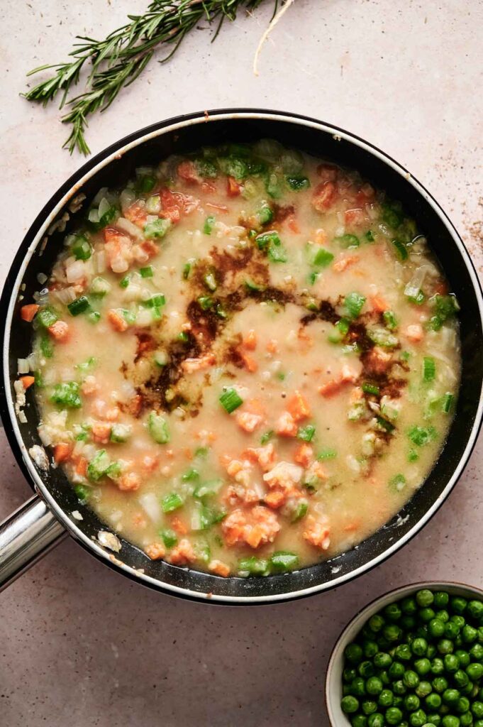 A frying pan with a mixture of diced vegetables and a creamy sauce evokes the comfort of a classic beef pot pie. Fresh rosemary is beside the pan, while a small bowl of peas rests nearby on a light-colored surface.