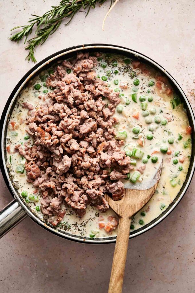 A skillet brimming with Beef Pot Pie filling—cooked ground meat, peas, and diced vegetables swathed in a creamy sauce. A wooden spoon rests inside the skillet, while fresh rosemary lingers nearby.