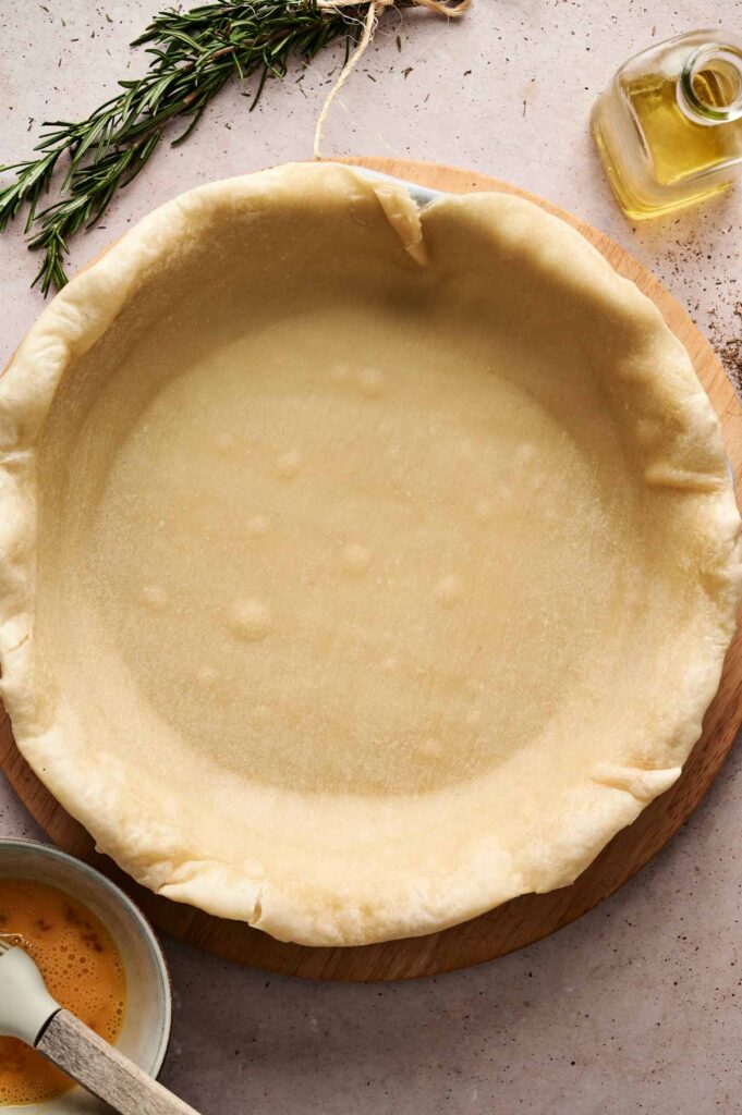 A hand uses a knife to cut dough on an unbaked beef pot pie crust, surrounded by a rosemary sprig, olive oil, and an egg wash bowl on the countertop.