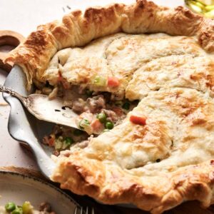 A golden-brown beef pot pie with a slice removed reveals a hearty filling of vegetables and tender meat. A serving utensil rests invitingly inside the pie dish.
