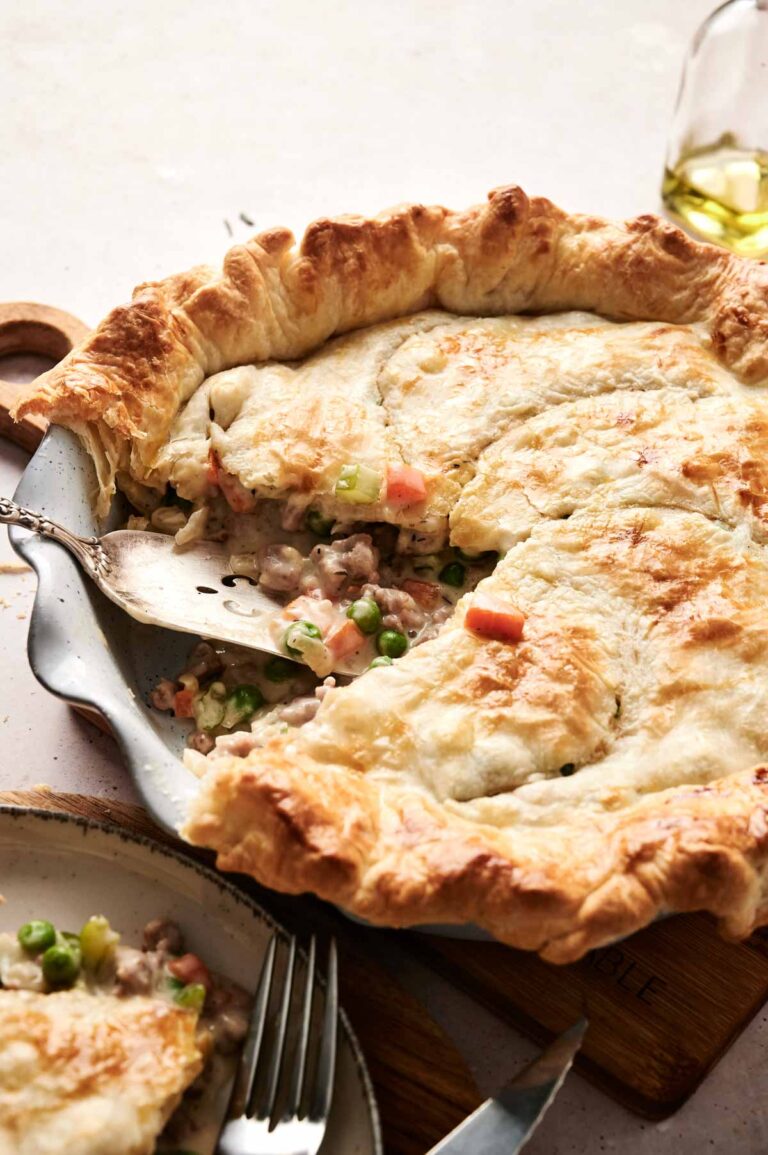 A golden-brown beef pot pie with a slice removed reveals a hearty filling of vegetables and tender meat. A serving utensil rests invitingly inside the pie dish.