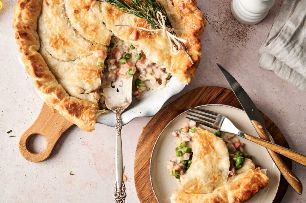 A savory beef pot pie with a flaky crust is partially served, revealing a hearty filling of mixed vegetables and tender meat. A portion rests on a plate next to cutlery on a wooden board.