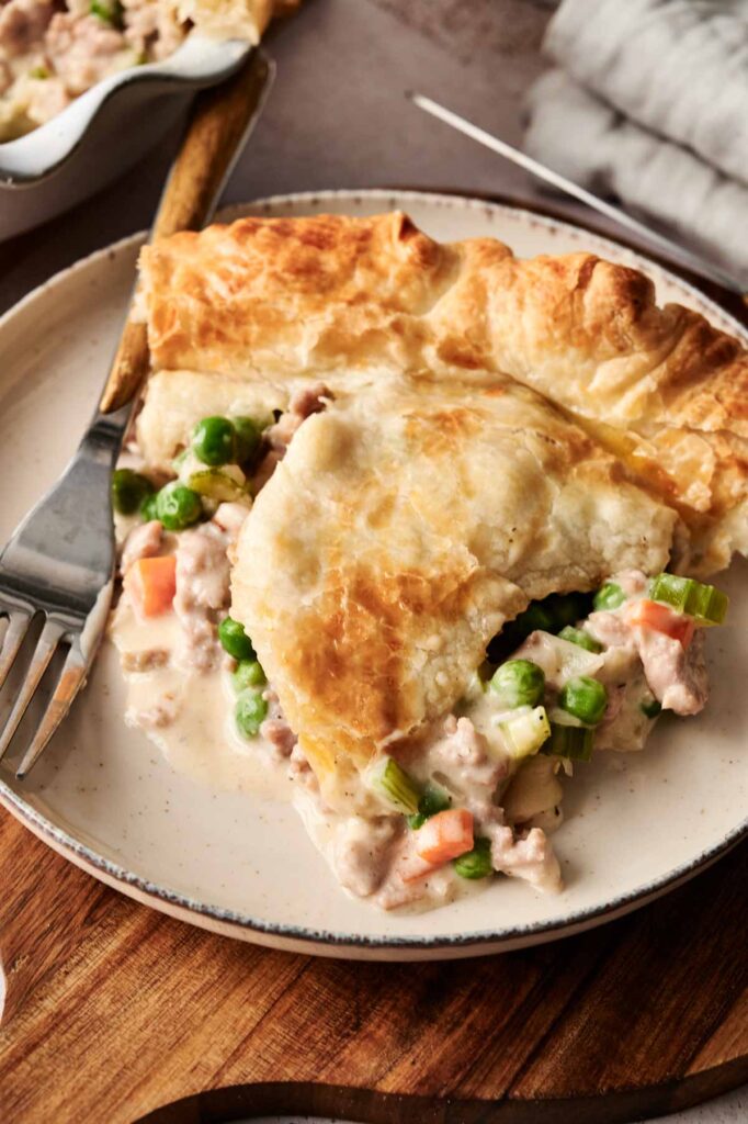 A slice of chicken pot pie sits on a plate, showcasing a flaky golden crust filled with savory chicken, peas, carrots, and creamy sauce. Reminiscent of its beef pot pie cousin, this dish is perfect comfort food. A fork rests beside the serving.