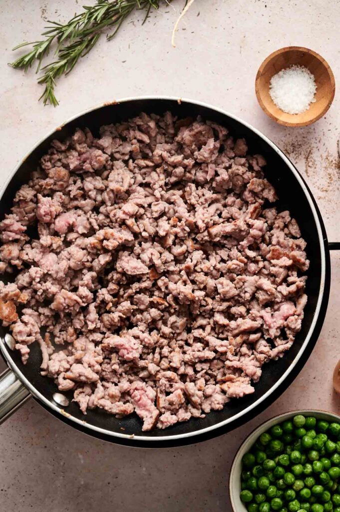 A frying pan filled with cooked ground meat sits ready to become the filling for a savory Beef Pot Pie. Nearby are a small bowl of salt, fresh rosemary sprigs, and a bowl of green peas, waiting to add flavor and texture.