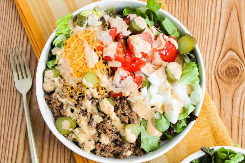 A bowl of Big Mac Salad with ground beef, shredded cheese, chopped tomatoes, pickles, onions, lettuce, and creamy dressing sits on a wooden table with a fork and napkin.