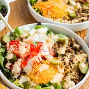 Salad bowls with chopped lettuce, ground beef, tomatoes, pickles, shredded cheese, and a drizzle of dressing on a wooden table.
