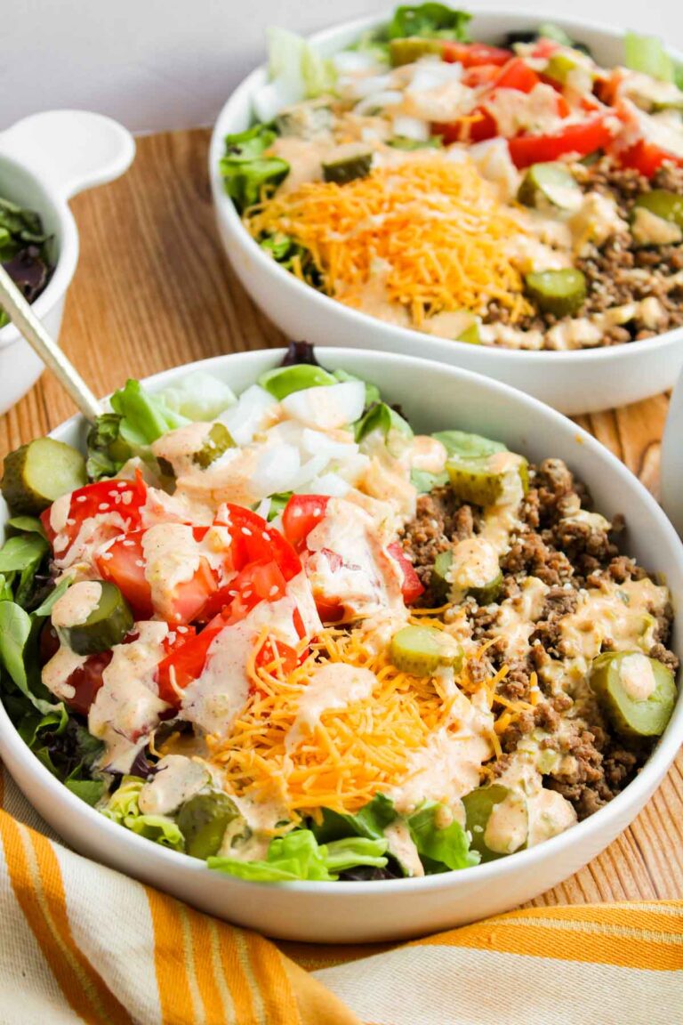 Salad bowls with chopped lettuce, ground beef, tomatoes, pickles, shredded cheese, and a drizzle of dressing on a wooden table.