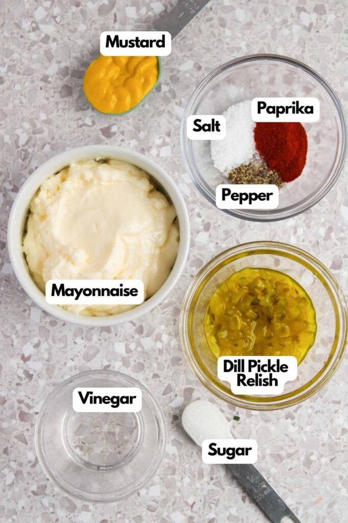 Ingredients for a sauce are arranged on a counter: mayonnaise, mustard, salt, paprika, pepper, dill pickle relish, vinegar, and sugar.