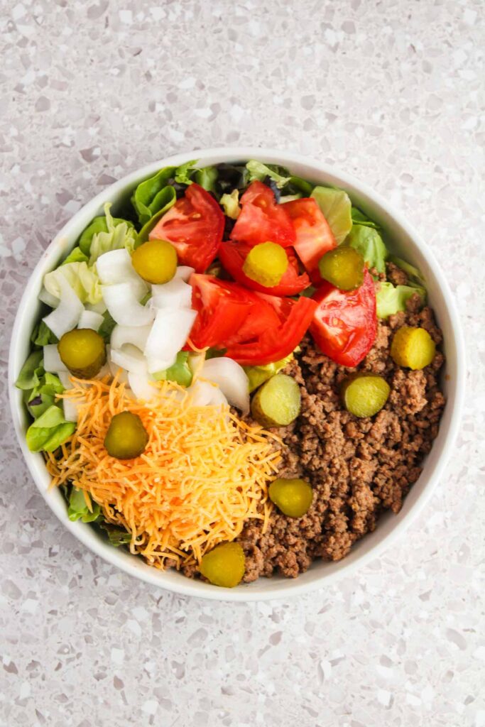 A bowl with ground beef, shredded cheese, chopped onions, lettuce, sliced tomatoes, and pickles on a speckled countertop.