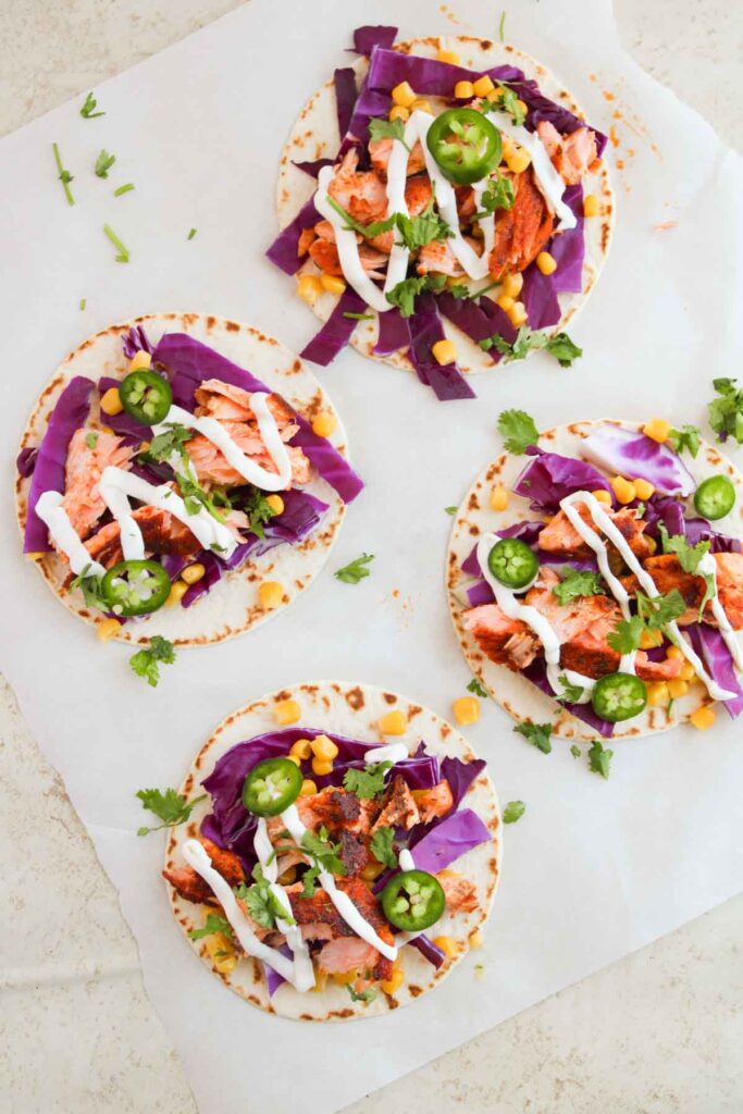 Four tacos on parchment paper, topped with purple cabbage, sliced jalapeños, corn, shredded meat, cilantro, and drizzled with white sauce.