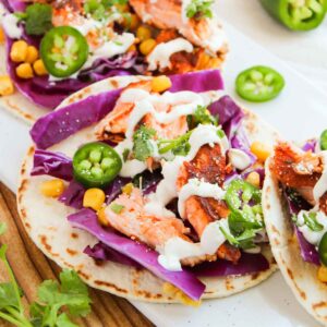 Three tacos with grilled salmon, purple cabbage, corn, jalapeño slices, and drizzled sauce on a white plate. A sliced jalapeño and cilantro garnish are nearby.